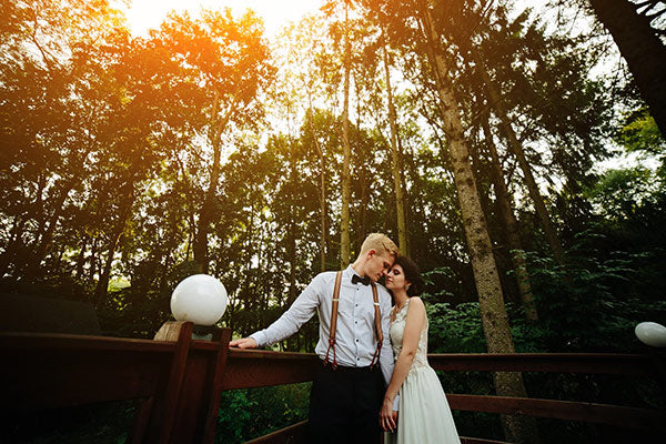 A bride and groom lean on a wooden fence in front of a forest at sunset.