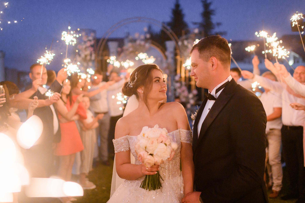 A bride and groom gaze at one another surrounded by wedding guests with sparklers at an outdoor wedding.