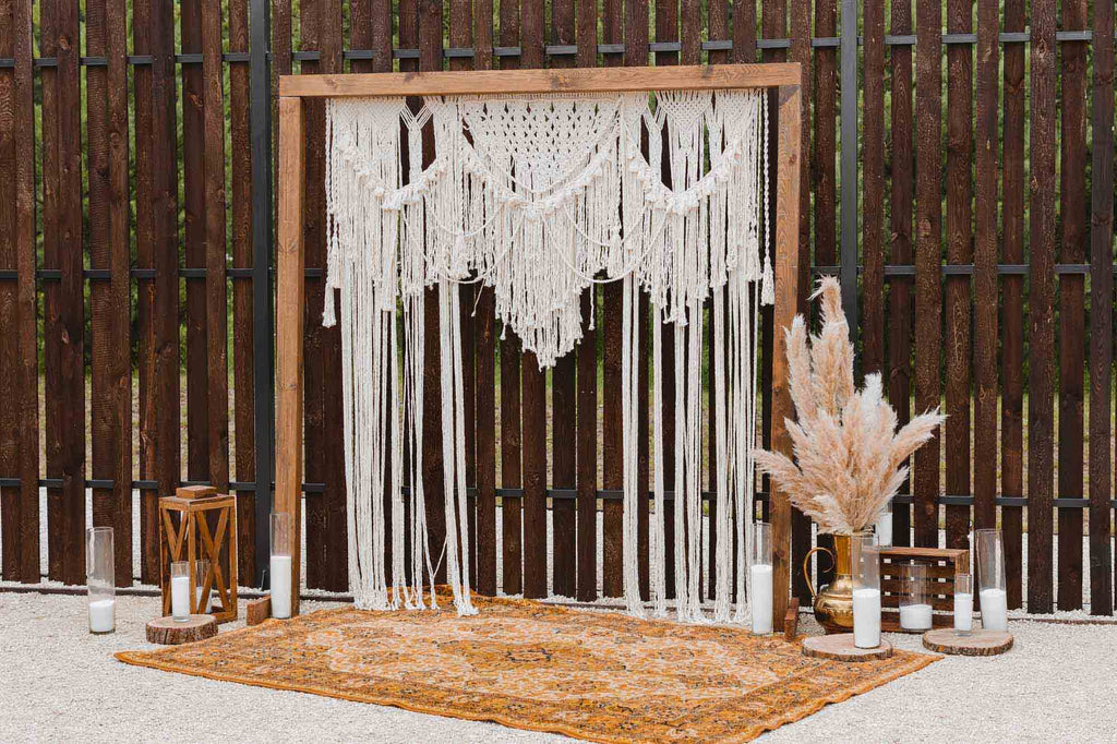 A wood frame wedding arch is fitted with white macrame curtains in front of wooden fence with white candles and fan foliage. 