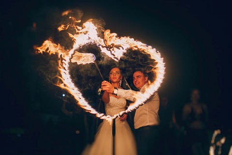 A bride and groom set a fire sculpture in the shape of a heart on fire at a nighttime outdoor wedding. 