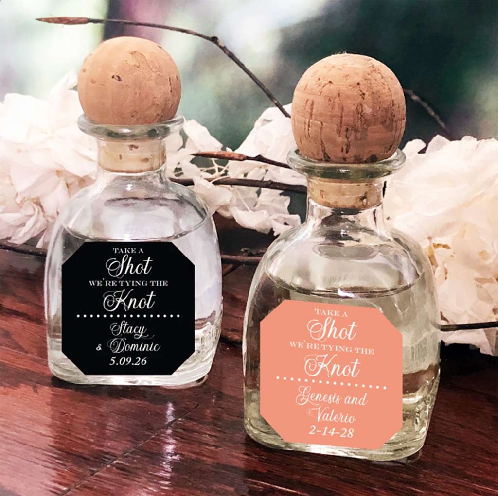 Two small mini patron bottle wedding favors rest on a wooden table imprinted with the poem, "Take a shot, we're tying the knot. Stacy & Dominic" followed by a wedding date. 