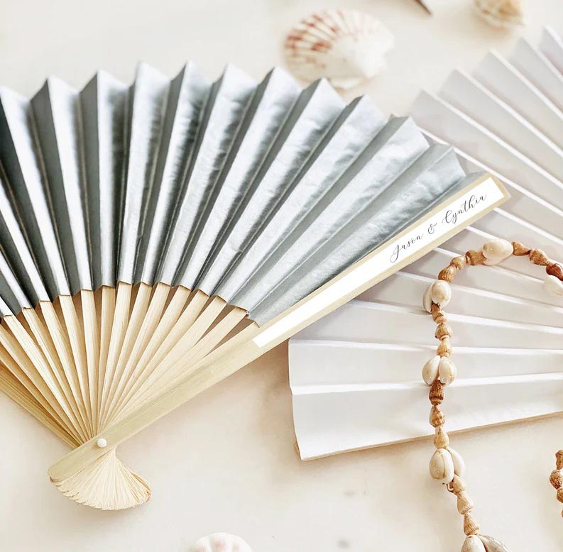 Two personalized wood and blue paper wedding fan favors are spread open on a white table.