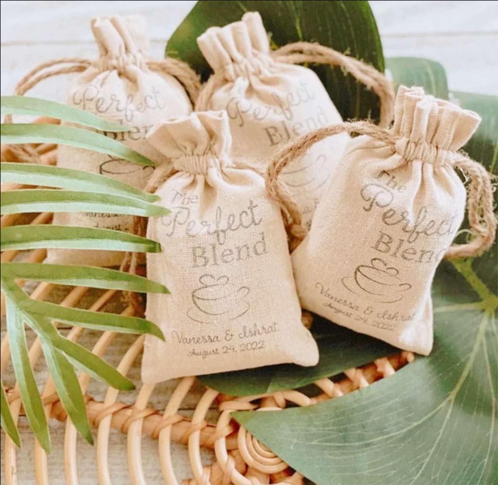 A wicker basket surrounded by leaves is filled with 3 personalized coffee bag wedding favors.