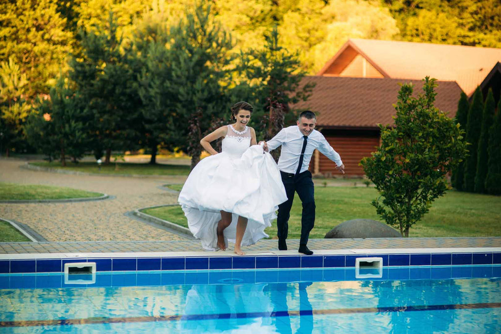 Young couple jumping in the swimming pool in a wedding suit and wedding dress at their poolside wedding.