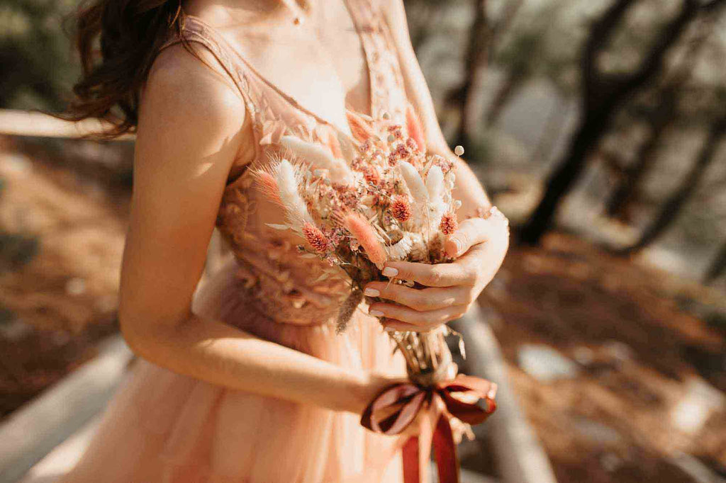 A boho bride in a pink sheer dress holds pink and white cattail bouquet to her chest.