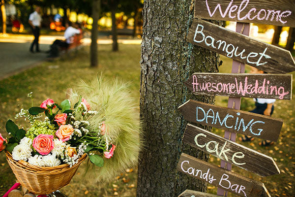A basket of flowers sits next to a sign comprised of several different arrows points wedding visitors to different wedding activities. A park setting is in the background. 