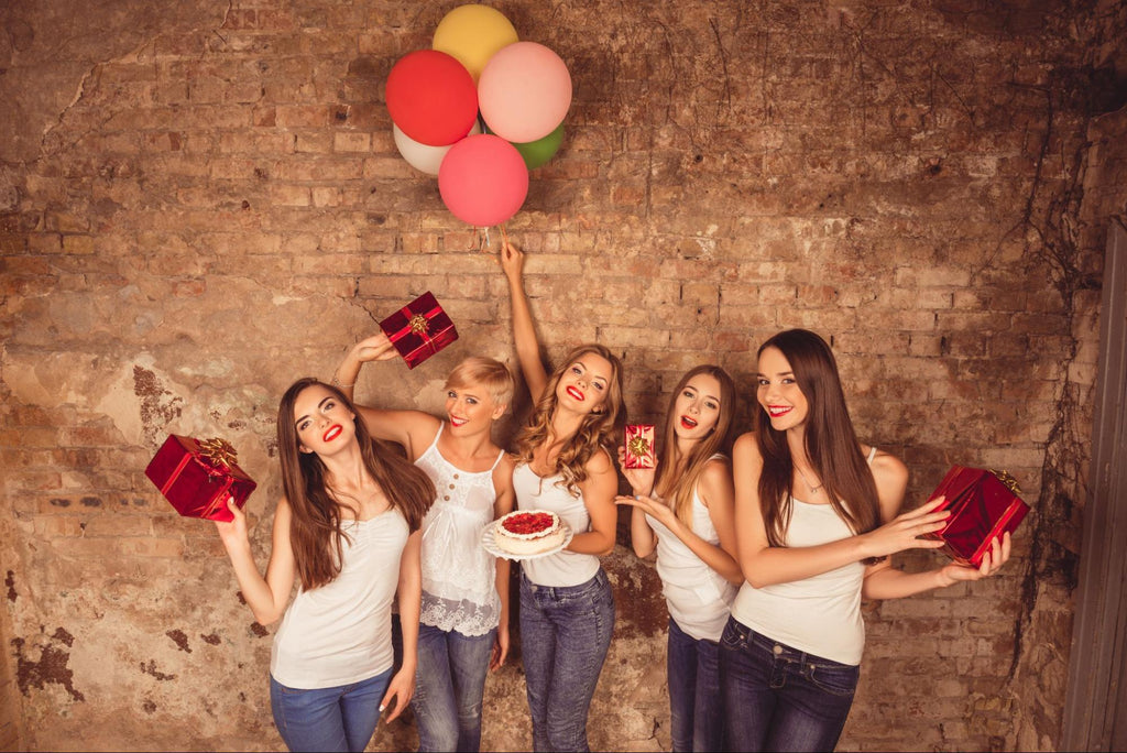 Five women are dressed in blue jeans and white tank tops. Four of the women hold gifts while the woman in the center holds a set of colorful balloons.