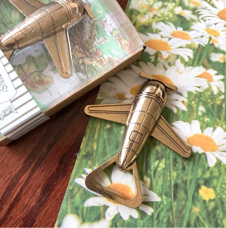 Two bonze airplane bottle openers destination wedding favors are sitting on a wood table. One bottle opener is packaged and the other  rest on a daisy print gift bag.