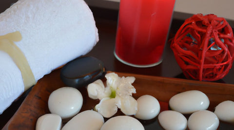 At home pamper party decorations featuring a white towel and wooden box filled with white decorative stones sitting next to a lit red candle and a small red wire sculpture in the shape of a ball.  