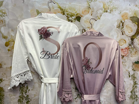 Two customized bathrobes are shown with intricate floral patterns on a single letter. One robe is white and reads bride. One robe is lavender and reads bridesmaid.
