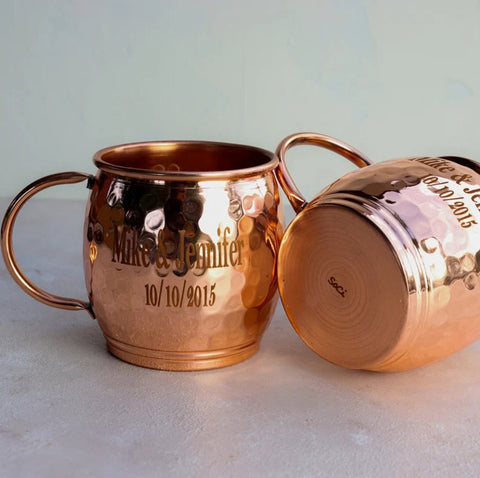 Two copper mug bohemian wedding favors rest on a grey table- one up right and one titled on the side - with the text "Mike & Jenny 10/10/2015" engraved on them.