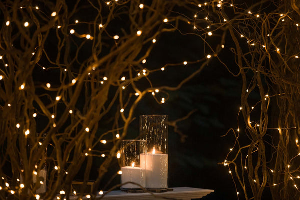 Two white candles in glass sit beside one another with many fairy lights in trees in the background of a nighttime wedding