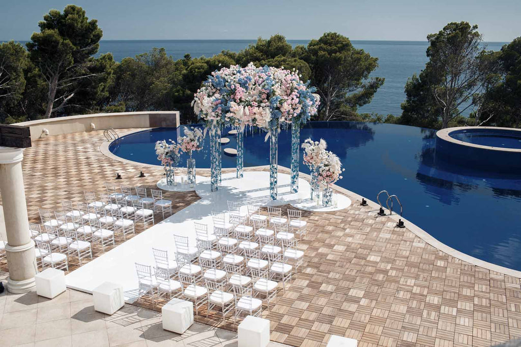 White chairs surround both sides of a white aisle leading to a dramatic flower arrangement of blue and pink flowers, elevated by four metal pillars in front of a swimming pool