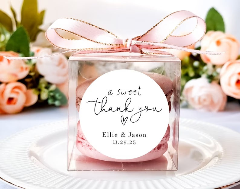 A pink and brown macaron are packed in a see through pink box with a pink bow and a label that reads, "A Sweet Thank You - Ellie & Jason" with a wedding date.