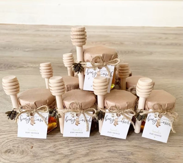 Eight small tan honey pots with matching wood honey dippers are tied shut with tan paper and tan twine. Each honey jar wedding favor has a small wedding couple tag.