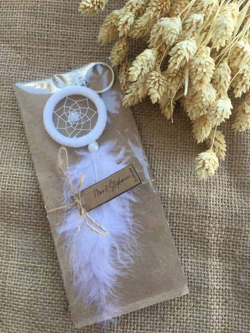 An all white dream catcher key chain with a long white feather is packaged with twine on a burlap table cloth. Wheat stalks rest beside it. 
