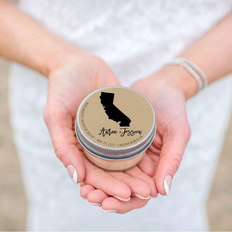 A woman holds a small glass wedding favor candle with a tin lid in her hands that features a customized label printed with the state of California and the names, "Anton & Jess" below it. 