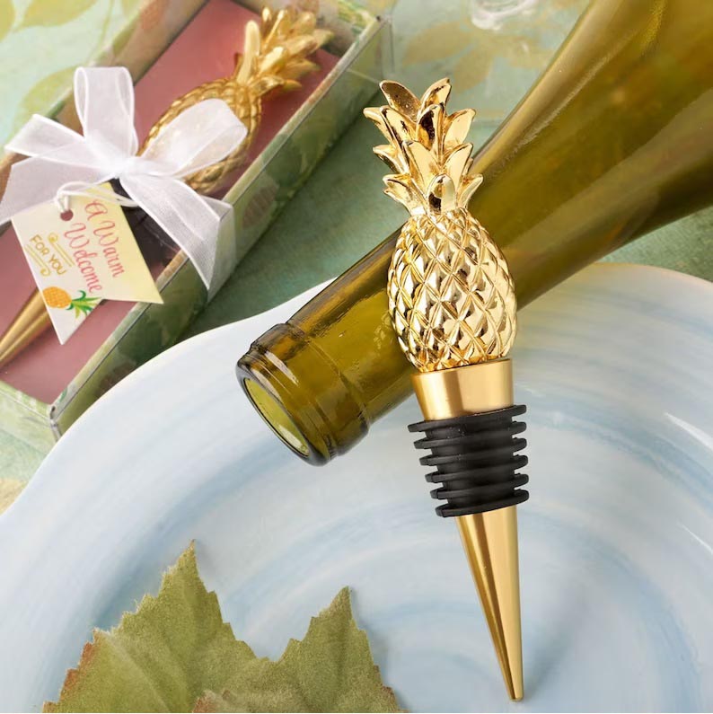 Two gold pineapple wine stopper destination wedding favors are sitting on a table. One wine opener is packaged and tied with a white bow. The other wine stopper is resting against the neck of a wine bottle on its side.