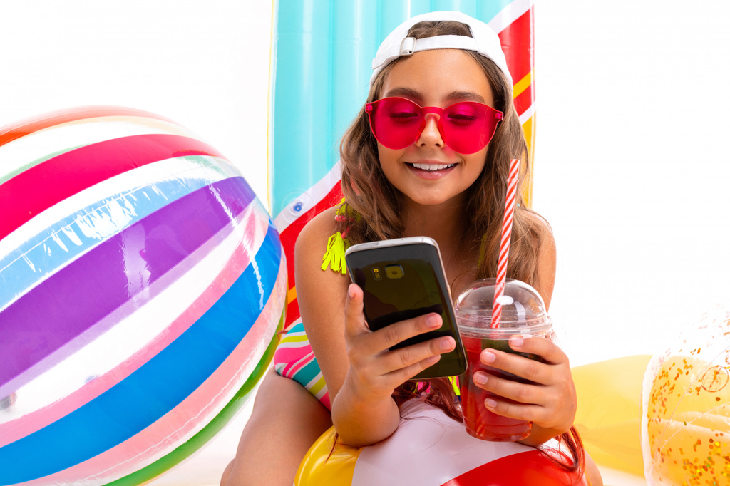 A woman checks her phone wearing a white hat and red sunglasses with a large beach ball pool party favor.