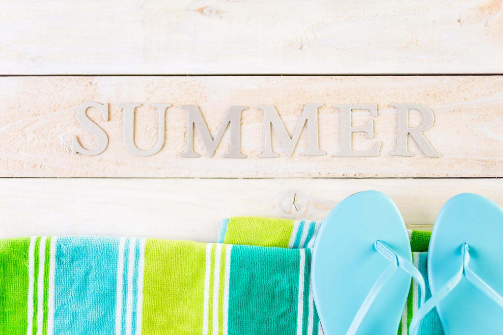 Light blue flip flops rest on top of blue and green striped towels on top of wood deck with the word "summer" written. 