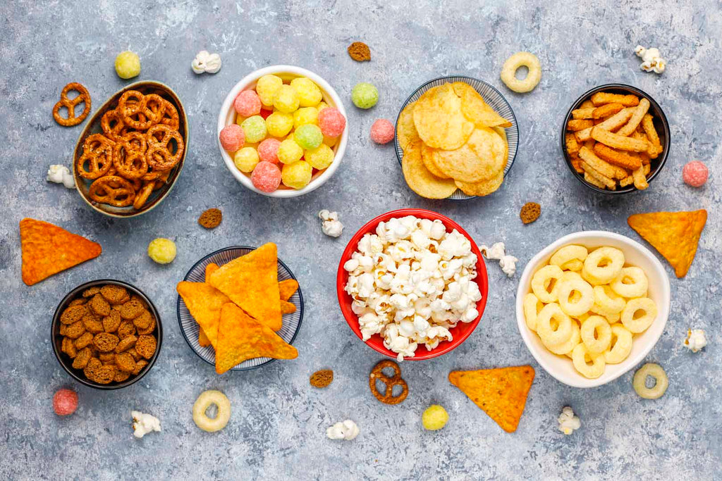A grey table is covered with bowls of pretzels, chips, crackers and popcorn.