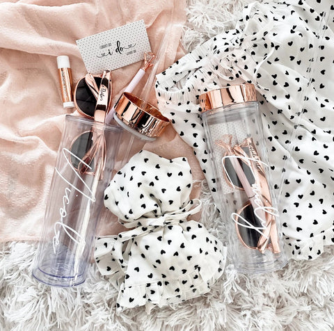 Two clear tumbler with rose gold lids and cursive font writing are filled with a pair of rose gold sunglasses and a rose gold lip balm. One tumbler rest on top of a white cloth gift bag with heart polka dots, while the other sits on a rose gold cloth. A closed polka dot gift bag is in between the two items.