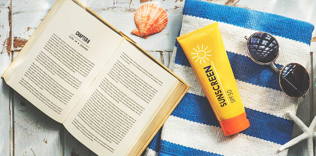 Suncreen sunglasses and an opened book rest on top of a blue and white beach towel. 