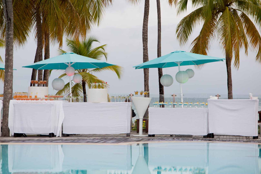 A pool is surrounded by white tables filled with orange cocktails, covered by blue patio umbrella pool decor for weddings .