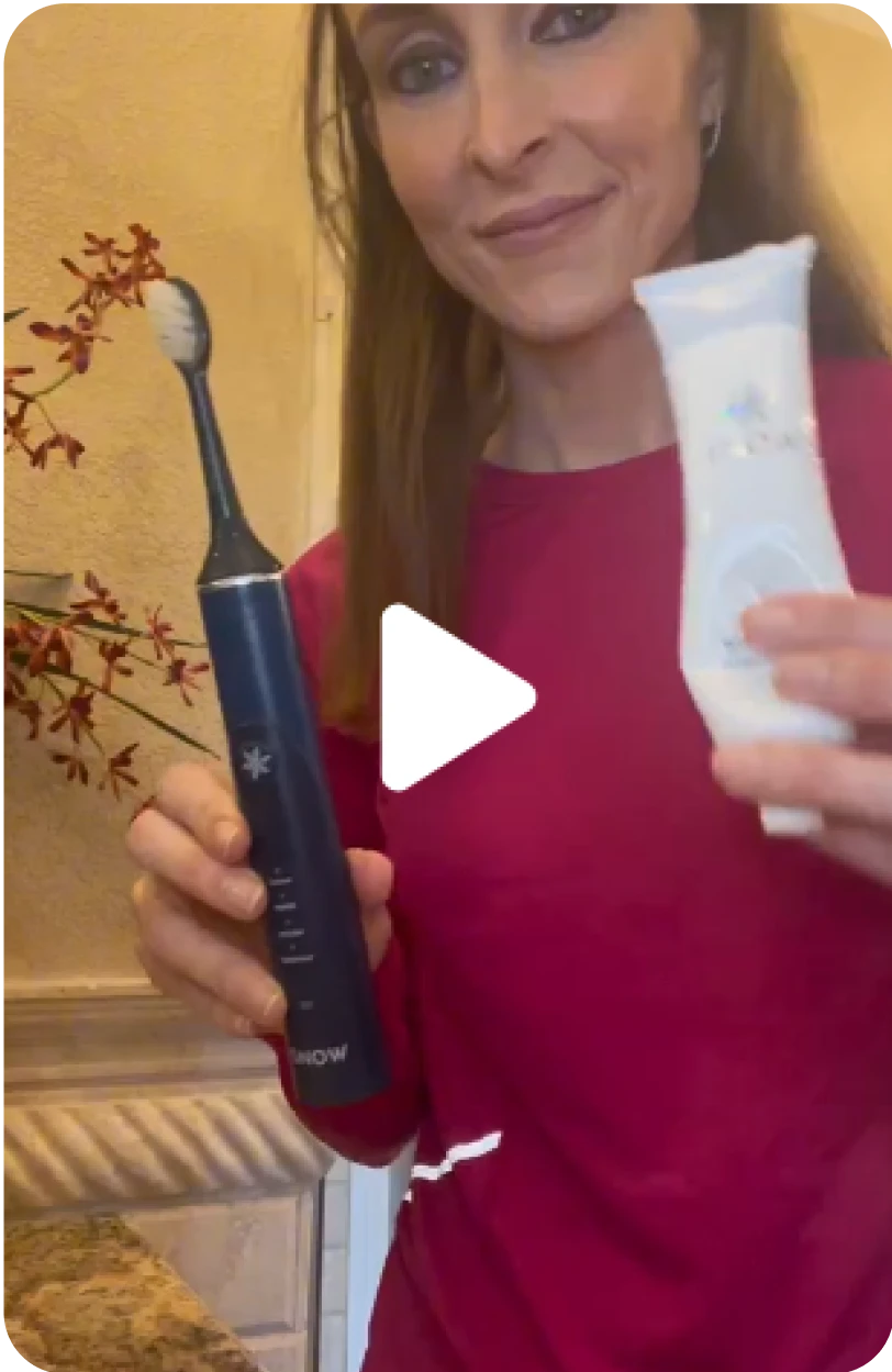 A woman holding an electric toothbrush and a tube of toothpaste in a bathroom.