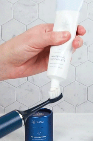 A hand applying whitening toothpaste to an electric toothbrush in a bathroom.