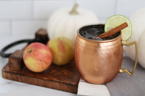 Apple cider mule cocktail in copper cup garnished with lime and cinnamon sticks with apples and pumpkin decor
