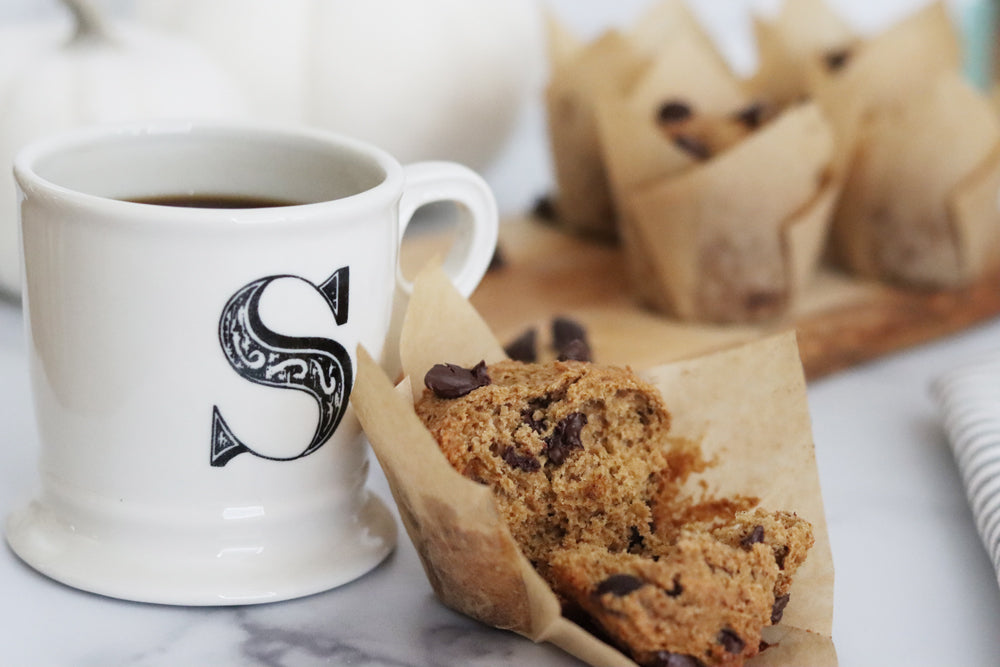 Mug of coffee with an S on it next to an unwrapped pumpkin butter chocolate chip muffin with more muffins in the background on a wooden cutting board
