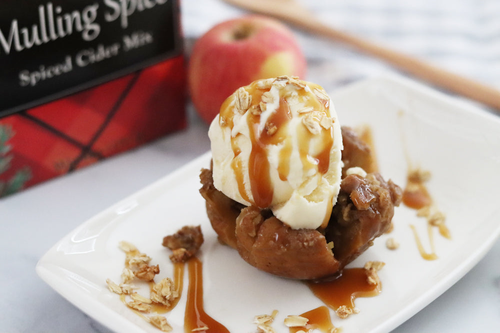 Baked apple topped with vanilla ice cream and caramel sauce next to fresh apples, cinnamon sticks, and Raven's Nest Mulling Spice Gift box