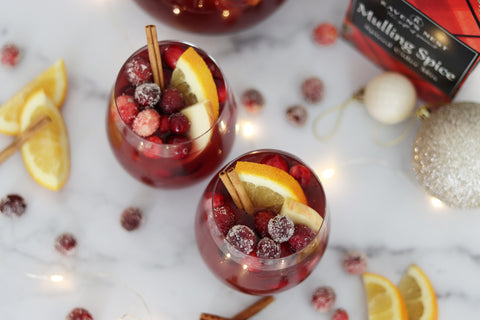 Two glasses of holiday sangria garnished with frosted cranberries, orange slices, and cinnamon sticks next to a box of Raven's Nest mulling spices