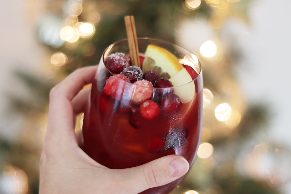 Woman holding glass of holiday sangria garnished with frosted cranberries, cinnamon sticks, and orange slices in front of the Christmas tree