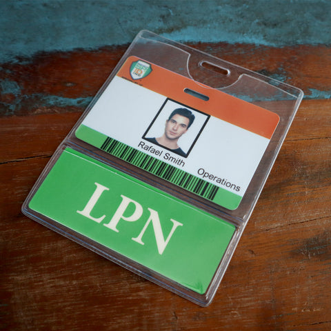 A Custom Printed BadgeBottoms® Horizontal (Badge Holder & Badge Buddy IN ONE) with a photo and the name "Rafael Smith, Operations" is displayed. The ID badge is encased in a clear holder along with a green card labeled "LPN." The background features a wooden surface partially painted blue.