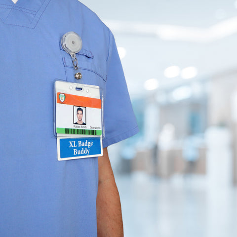 Close-up of a person in blue scrubs wearing a name badge labeled "Peter Smith, Operations" and an Oversized Custom Printed Horizontal XL Badge Buddy (Extra Large Size) below it. The background is blurred, suggesting an indoor setting, possibly a hospital or medical facility.