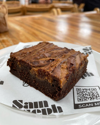 A chocolate brownie, served on a place with branded greaseproof paper. The paper has a black and white logo printed across it and features a QR code