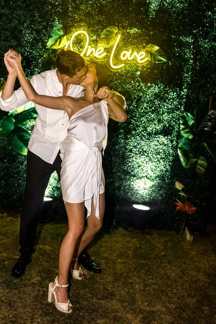 The couple kissing in front of a neon sign that says "One Love".