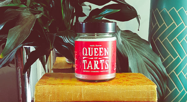 Queen of Tarts candle on a yellow book with green plants behind it