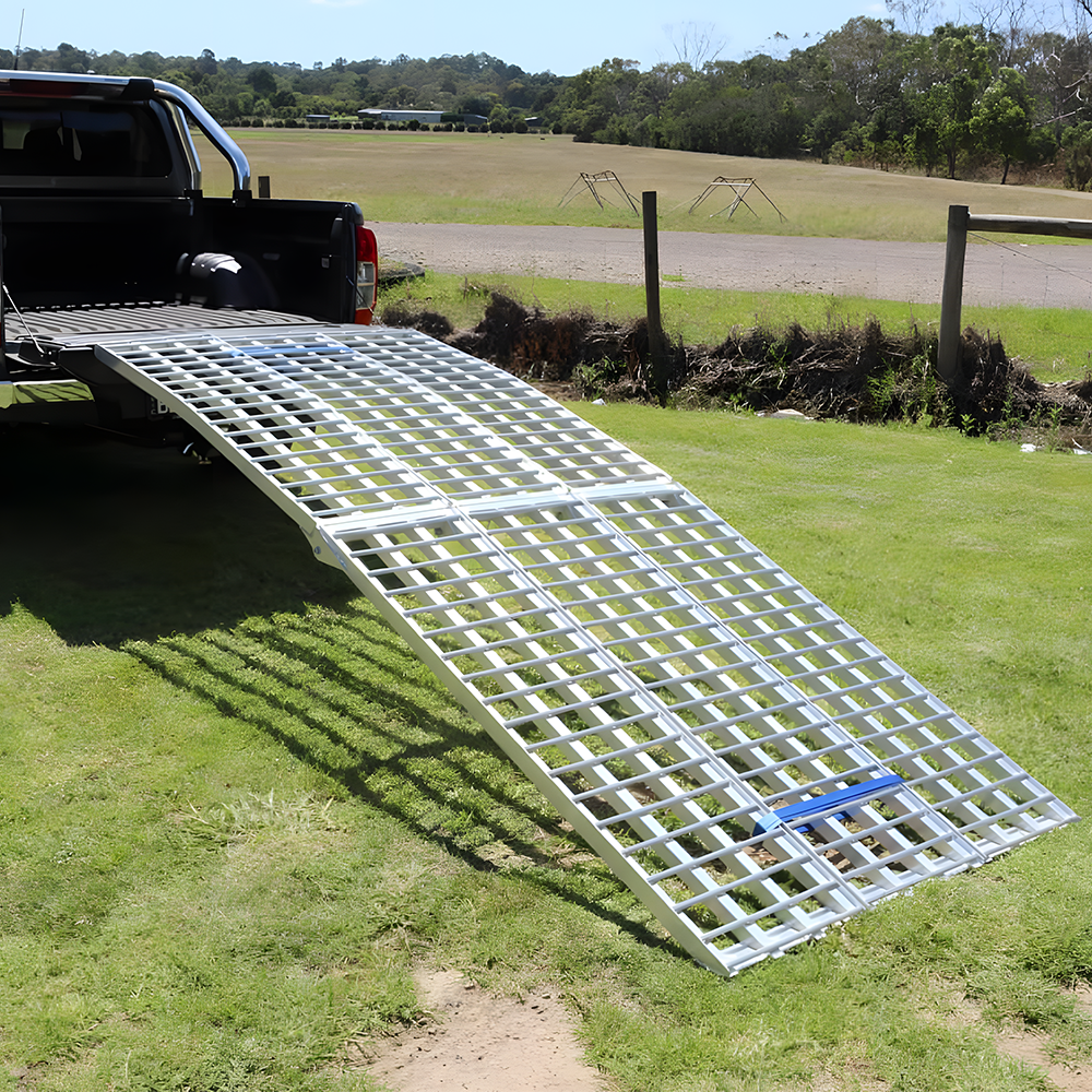 Aluminium loading ramp secured to the tailgate of a pickup truck, curving down to the grassy ground for easy vehicle loading.