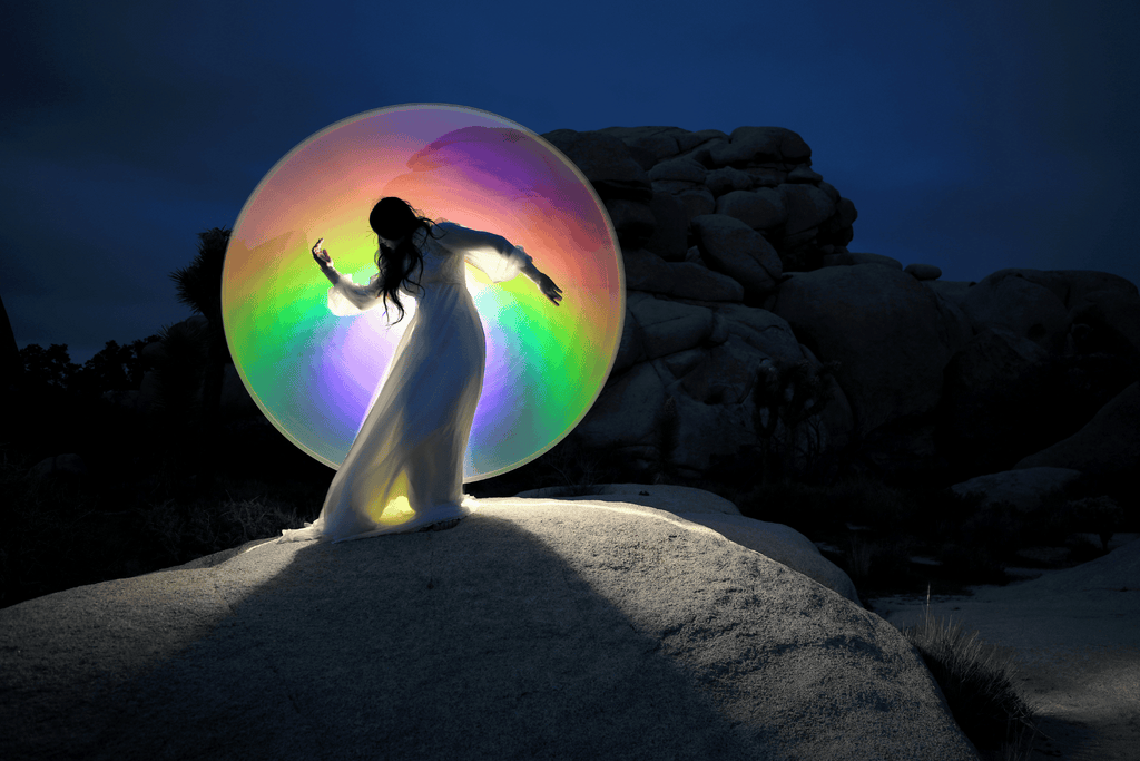 light painting photo of a woman in white dress standing on a rock behind a colorful round orb