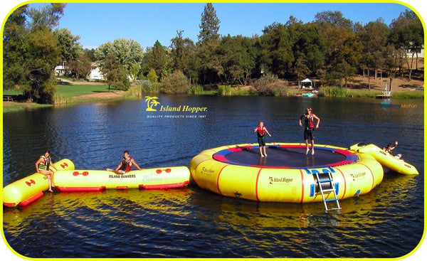 Island Hopper Giant Jump 25ft Water Trampoline Park. 2 young boys bounce on the lake trampoline while 2 others sit and play on the pad and the water attachments.