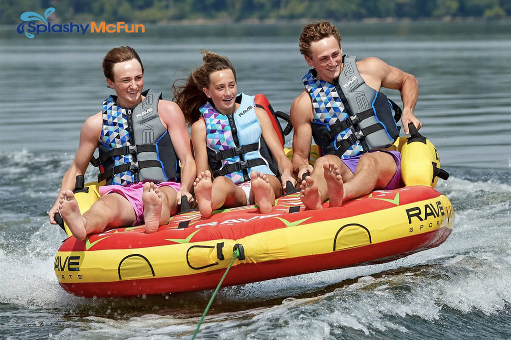 This is the RAVE #Epic 3-Person Towable Boat Tube with 3 people sitting on it, holding onto the handles while being dragged by a speedboat on the water.