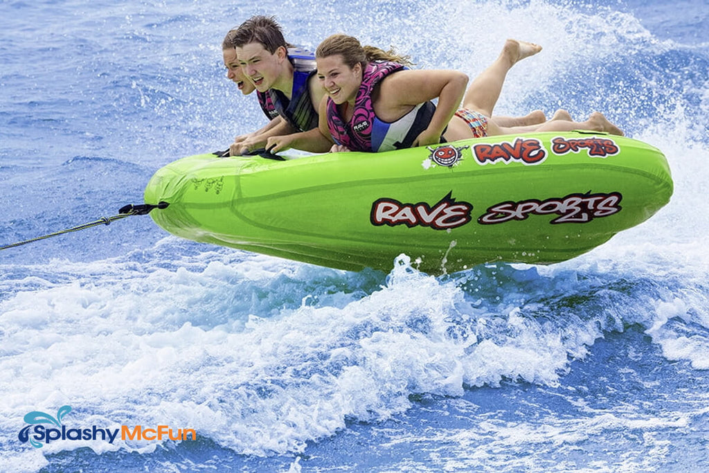 This is the Rave Mambo 3-Person Towable Tube with 3 people laying chest-flat on it, holding onto the handles while being dragged by a speedboat on the water.