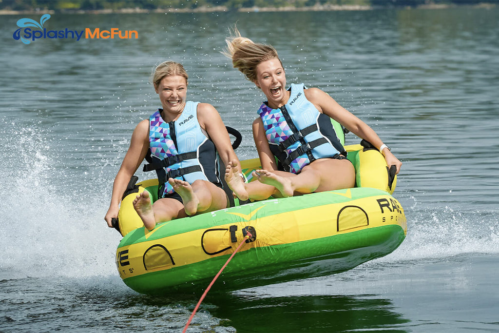 This is the Rave #Stoked 2-Person Towable Boat Tube with 2 people sitting on it in, holding onto the handles while being dragged by a speedboat on the water.