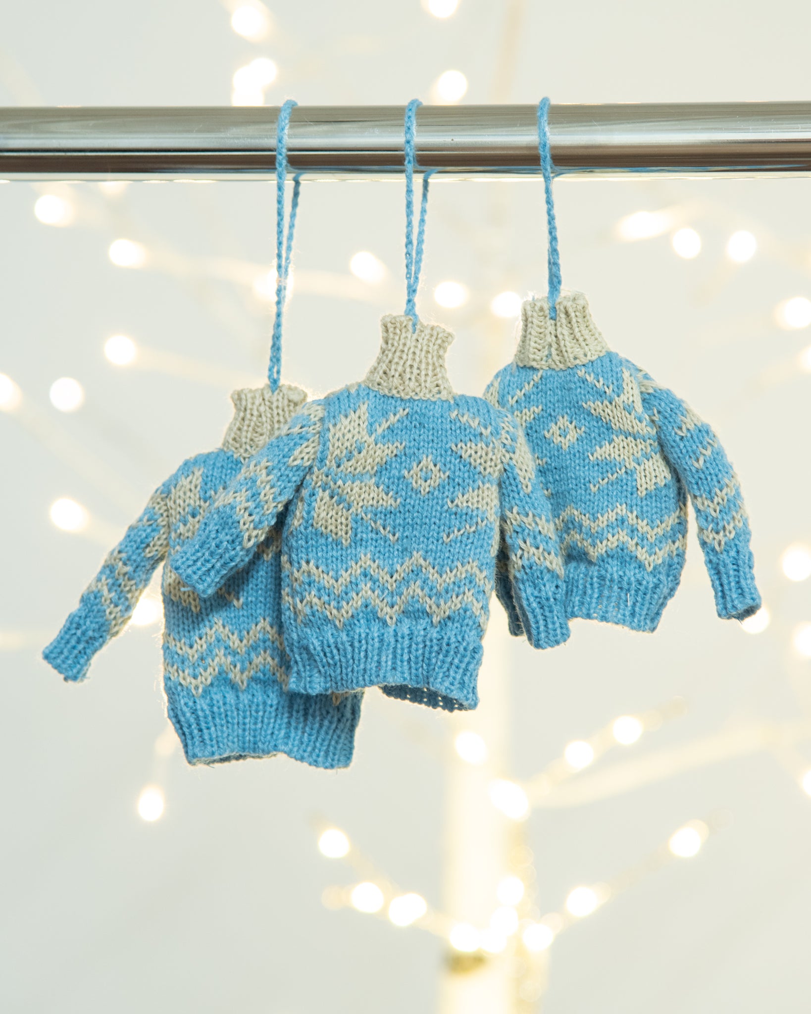 three mini sweater ornaments with snowflake pattern hanging on a rack in front of a white birch tree with tree lights