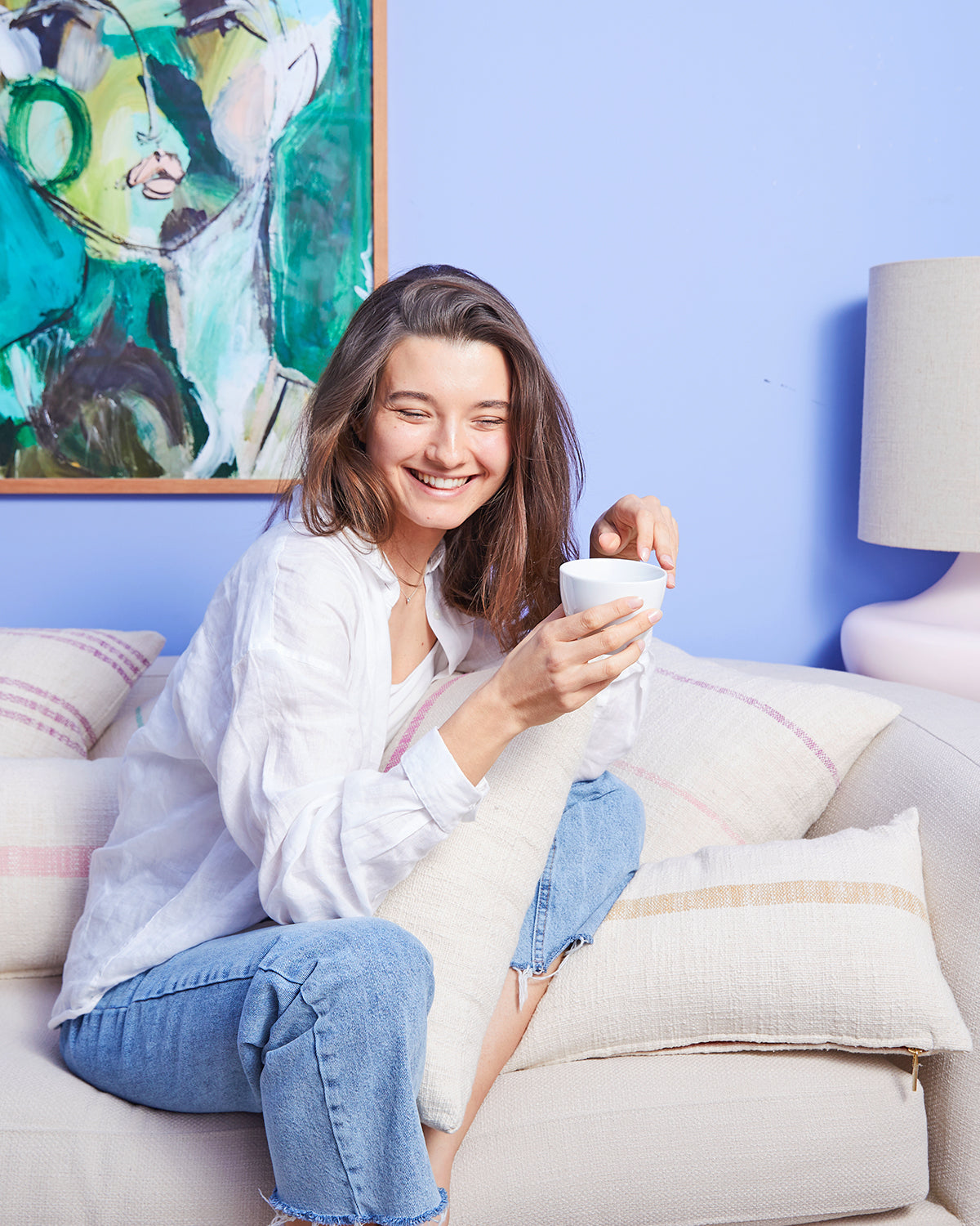 Woman laughing holding cup of coffee on a couch filled with pillows
