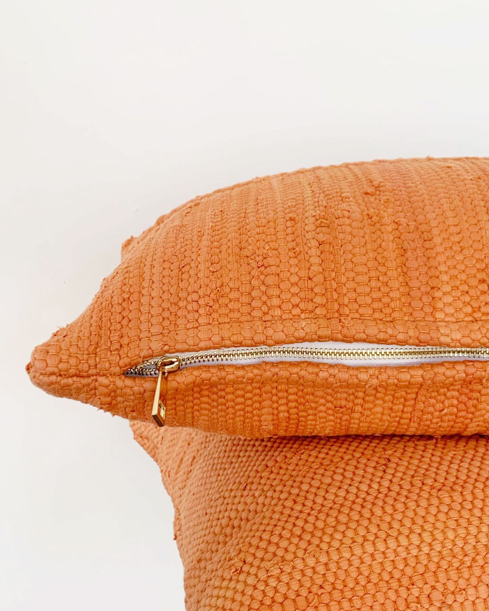 two orange pillows stacked, close up of gold zipper detail. In front of white wall