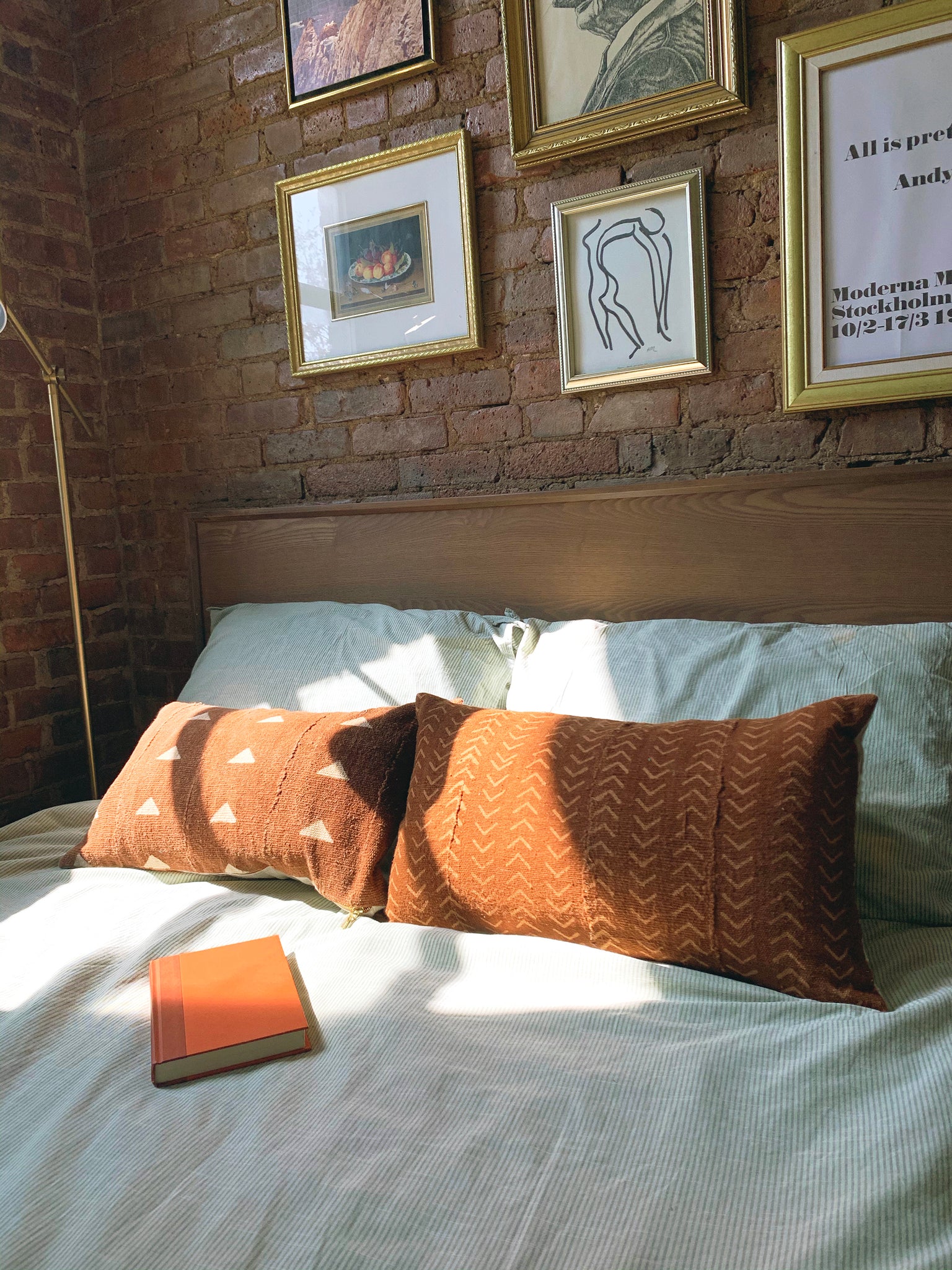 A closeup of a bed in front of a brick wall with gold picture frames. two long rust colored mud cloth pillows and an orange book on top of bed.